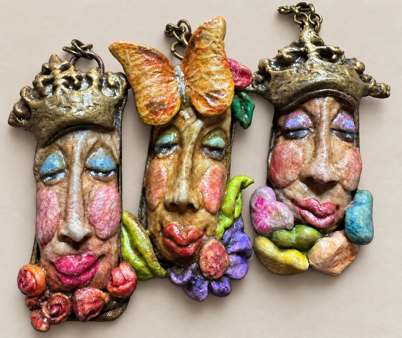 Three sculpted face pendants with crowns, a butterfly and colorful floral details, resting on a warm neutral surface
